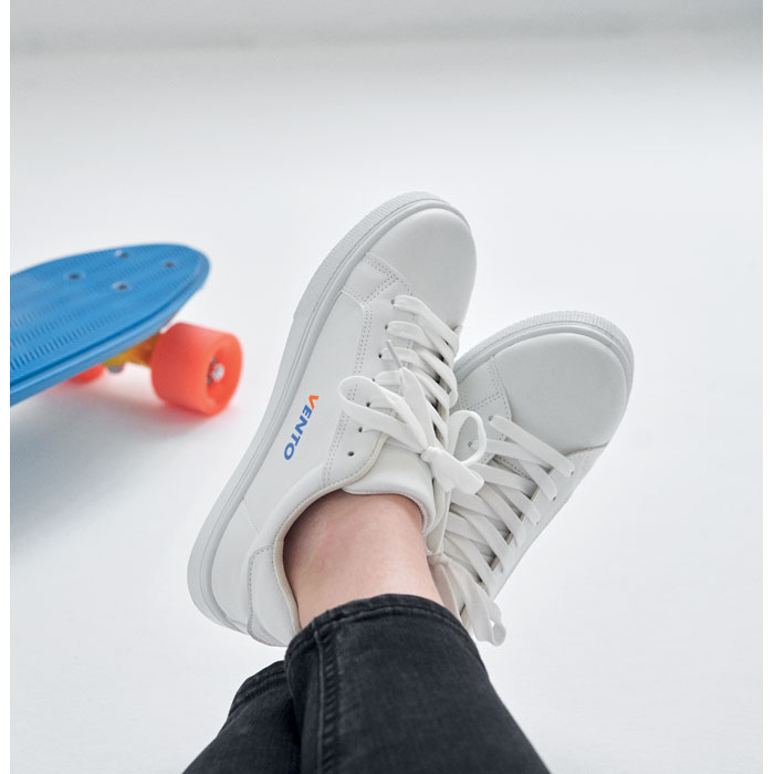 Zapatillas blancas Vento junto a un skate azul con ruedas naranjas sobre fondo blanco